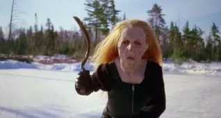 A screen shot taken from the 1983 horror film Curtains. It shows a woman in an old lady mask brandishing a scythe.