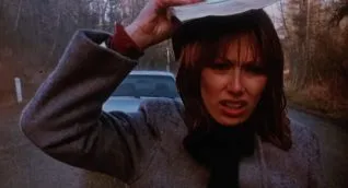 A screen shot taken from the 1983 horror film Curtains. It shows a woman standing on a road in the rain.