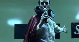 A screenshot taken from the 2004 horror film Calvaire. It shows a man in a cape singing.