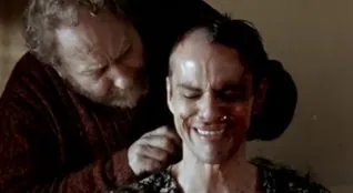 A screenshot taken from the 2004 film Calvaire. It shows a man crying in a dress, while another shaves his head.