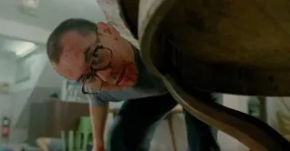 A screen shot taken from the 1993 horror film, The Untold Story. It shows a bloodied man peering underneath a table while brandishing a large knife.